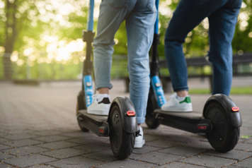 Two people on e-scooters. They each have a foot on an e-scooter and the other foot flat on the ground