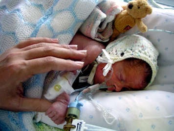 Premature baby in a crocheted bonnet sleeping in incubator with tubes connected to their nose and arm. An adult's hand rests on the baby's side