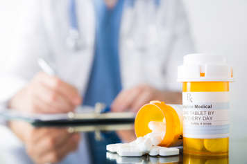Two prescription medication bottles on a doctor's desk. The doctor sits at the desk and writes on a notepad in the background