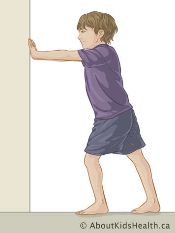 Child standing facing a wall with both hands on the wall with one leg stretched behind them and one stepped toward the wall