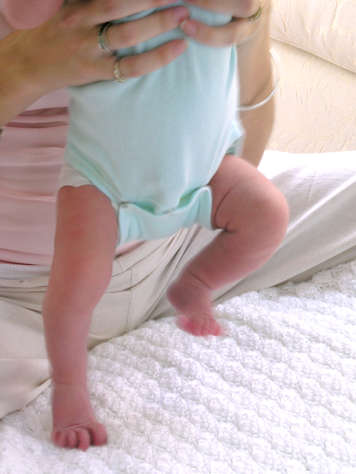 Newborn standing on one foot, with the other leg bent at the knee, while being held at the waist