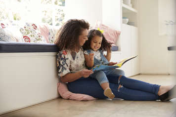 Woman sitting on the floor with a toddler sitting on her lap, holding a book open in front of them