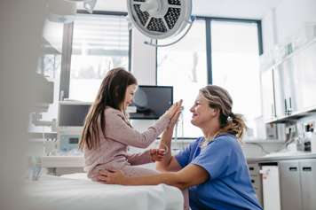 Child and health-care provider high-fiving. Child sits on hospital bed and health-care provider crouches in front of her