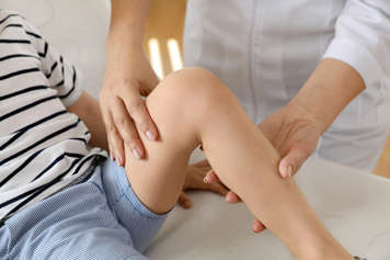 Closeup on a child's leg, which they have bent at the knee while a doctor holds one hand to the child's thigh and the other to the calf